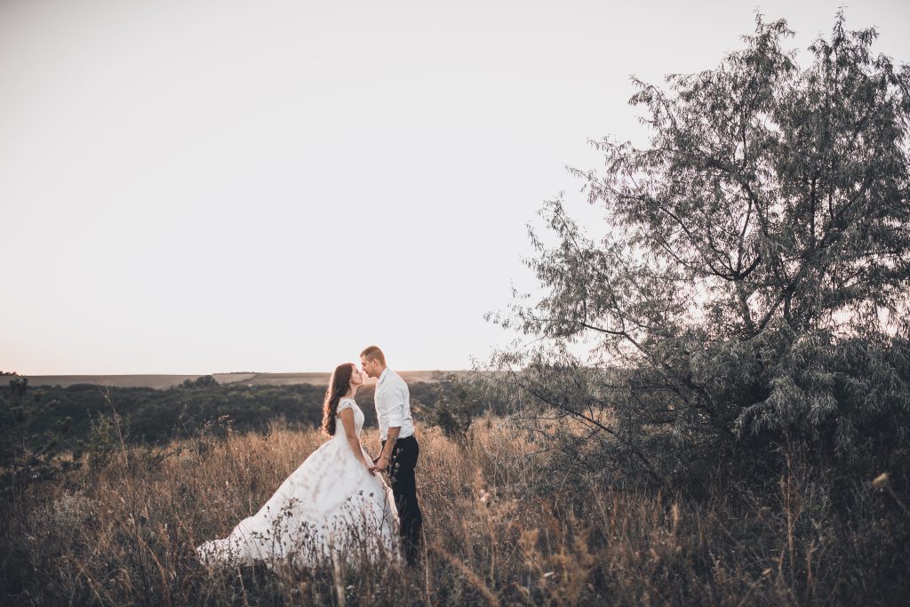 Bride and groom standing close together in a grassy field at sunset, sharing an intimate moment beside a tree in a natural outdoor setting.