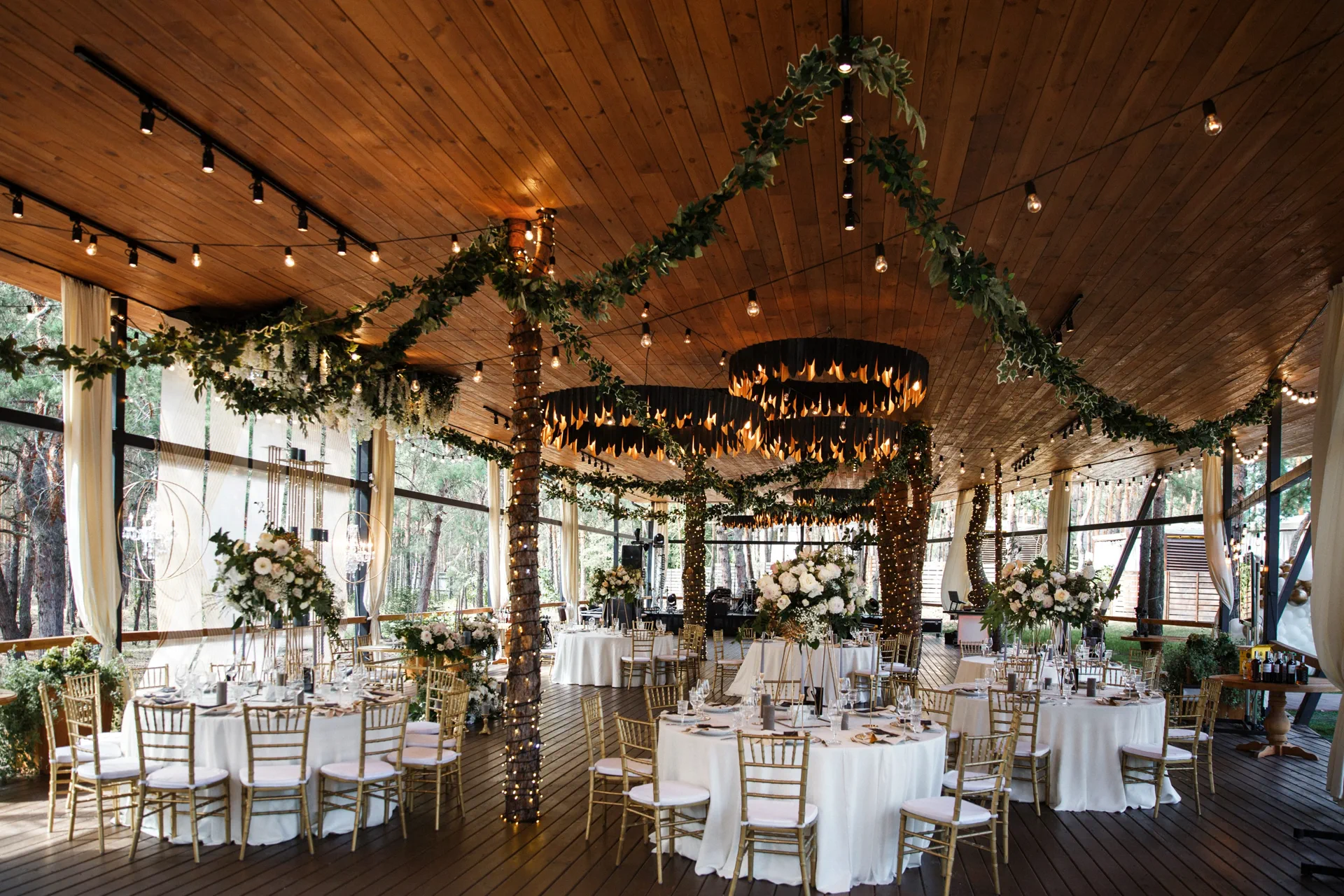 Elegant indoor wedding reception with round tables, white linens, tall floral centerpieces, and warm string lights and greenery draped across a wooden ceiling.