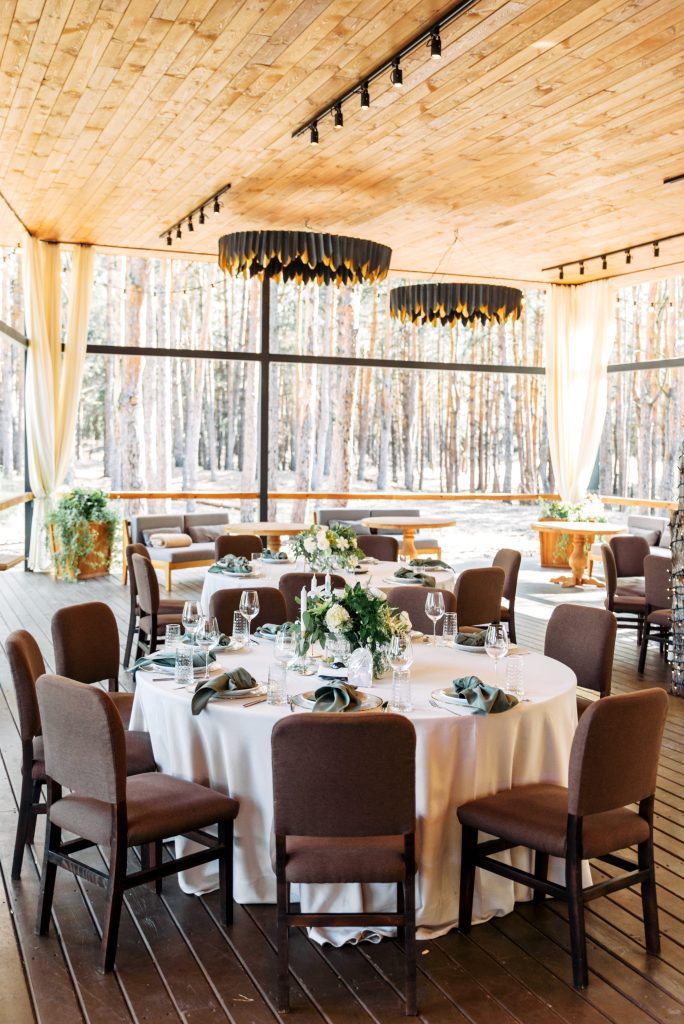 Elegant indoor event space with round tables set for dining, floral centerpieces, and soft lighting, overlooking a forest through large windows.