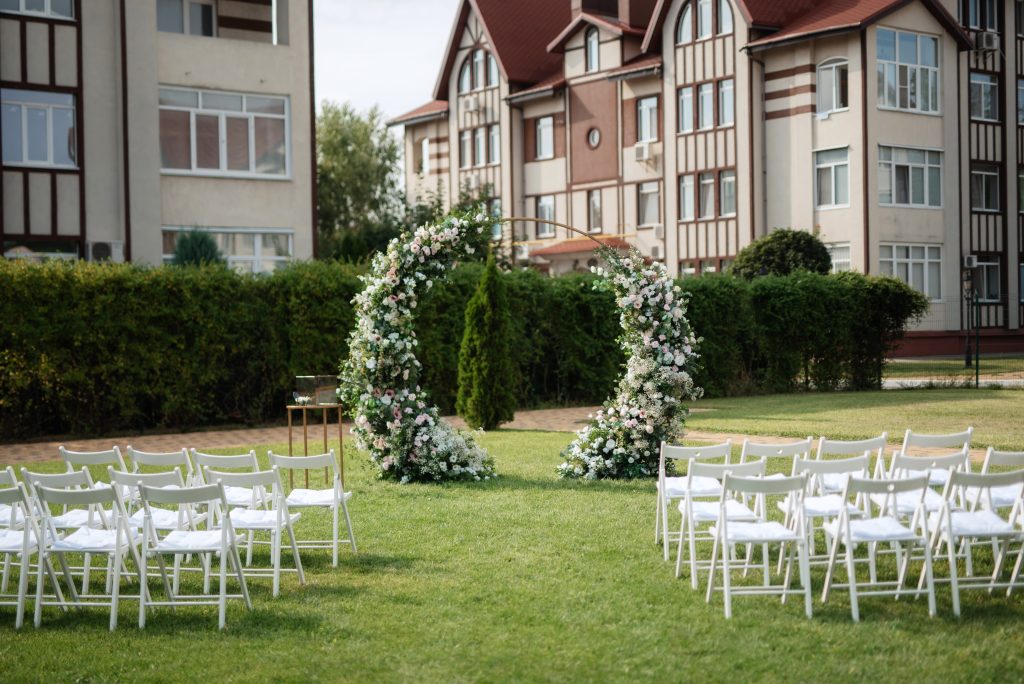 Outdoor wedding ceremony setup with white chairs arranged on a lawn facing a floral arch, set against a backdrop of residential buildings and greenery.