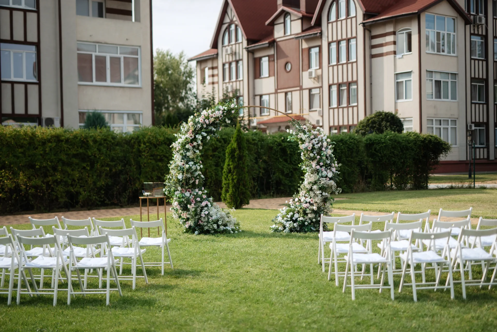 Outdoor wedding ceremony setup with white chairs arranged on a lawn facing a floral arch, set against a backdrop of residential buildings and greenery.